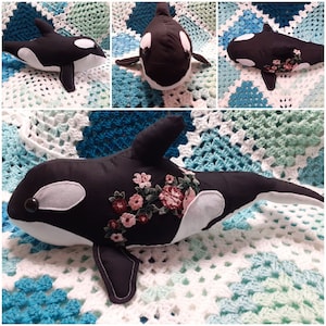 May include: Black and white plush orca with embroidered pink and green flowers. The orca is lying on a white and blue crocheted blanket.
