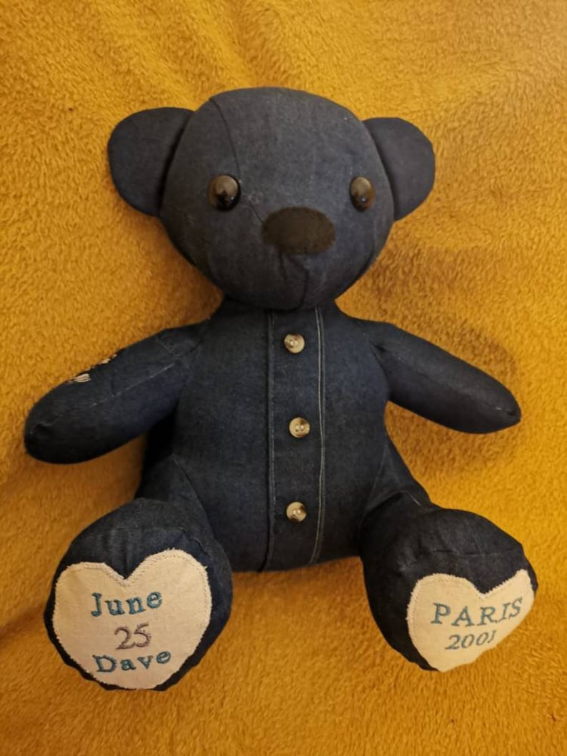 Pu&ograve; includere: Un orsacchiotto di jeans blu con occhi neri e naso nero. L'orso indossa una camicia di jeans con bottoni. I piedi dell'orso hanno cuori ricamati con il testo "June 25 Dave" e "Paris 2001".