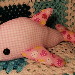 May include: Pink and white gingham dolphin plush toy with pink and yellow heart patterned fabric on the fins. The toy is lying on a crocheted blanket.