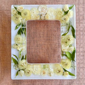 May include: A white square picture frame with a border of pressed white flowers and greenery. The frame is made of clear resin and has a rustic, natural look.