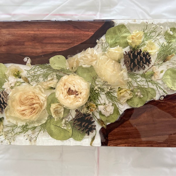 Può includere: Un vassoio rettangolare in legno con un top in resina trasparente, che presenta fiori pressati e pigne. I fiori sono bianchi e crema, con foglie e steli verdi. La resina è trasparente e lucida, e il legno è di un colore marrone intenso.
