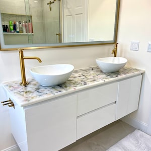 Könnte beinhalten: Ein weißer Badezimmer-Waschtisch mit zwei weißen Porzellan-Waschbecken und goldenen Armaturen. Der Waschtisch hat eine weiße Arbeitsplatte mit einem Blumenmuster und zwei Schubladen. Der Waschtisch ist an der Wand montiert.
