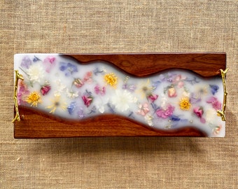 May include: A rectangular serving tray made of wood with a white resin inlay. The resin is filled with pressed flowers in shades of pink, purple, white, and yellow. The tray has two gold metal handles.