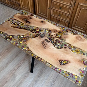 Puede incluir: Una mesa de madera con un acabado de resina epoxi transparente y flores secas incrustadas. La mesa es de color marrón claro con un patrón de veta de madera natural. Las flores son de varios colores, incluyendo amarillo, blanco, rosa y morado. La mesa está soportada por una base de metal negro.