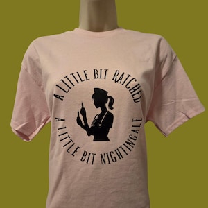 May include: A light pink t-shirt featuring a black silhouette of a nurse holding a syringe, surrounded by the text "A LITTLE BIT RATCHED, A LITTLE BIT NIGHTINGALE" in a circular design.