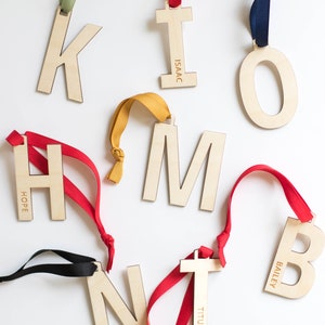 May include: Wooden letter ornaments with colorful ribbons. The letters K, I, O, H, M, N, T, and B are displayed, some with names engraved. Ribbons are in green, red, blue, yellow, and black.