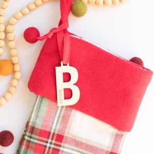 Puede incluir: Un calcetín navideño rojo con un estampado de cuadros, una etiqueta de madera con la letra "B" y una cinta roja. La etiqueta está grabada con el nombre "Bailey". Bolas de fieltro decorativas y cuentas de madera están esparcidas alrededor.
