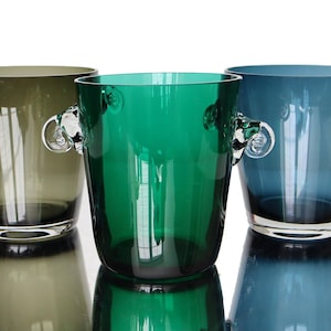 May include: Three colored glass buckets with handles. The buckets are in shades of green, teal, and blue. Each bucket has a clear glass handle on either side. The buckets are sitting on a reflective surface.