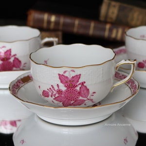May include: A set of three white porcelain teacups and saucers with a pink floral pattern and gold trim. The cups have a scalloped rim and a delicate floral design. The saucers have a matching floral design and a gold rim.