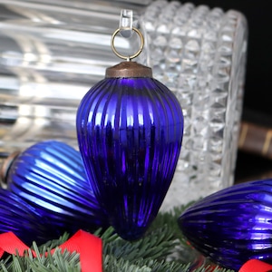 Könnte beinhalten: Ein Satz von vier blauen Weihnachtskugeln aus Glas mit gerippter Oberfläche. Die Kugeln sind auf einem Bett aus grünen Kiefernästen angeordnet.