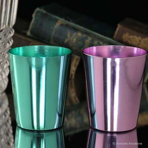 K&ouml;nnte beinhalten: Zwei kleine Metallbecher, einer in T&uuml;rkisgr&uuml;n und einer in Hellrosa, mit gl&auml;nzendem Metallic-Finish. Die Becher stehen auf einer reflektierenden Oberfl&auml;che.