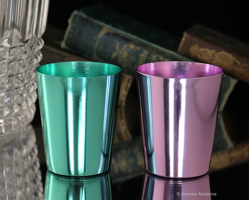 K&ouml;nnte beinhalten: Zwei kleine Metallbecher, einer in T&uuml;rkisgr&uuml;n und einer in Hellrosa, mit gl&auml;nzendem Metallic-Finish. Die Becher stehen auf einer reflektierenden Oberfl&auml;che.