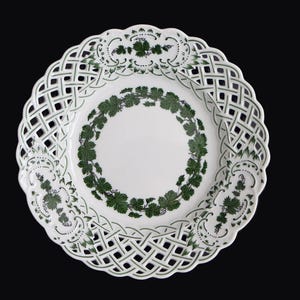 Può includere: Piatto in ceramica bianca con bordo a griglia e motivo a foglie verdi. Il piatto presenta un anello centrale di foglie e ulteriori motivi a foglie sul bordo, creando un'estetica decorativa ed elegante. Il piatto è posto su uno sfondo nero.