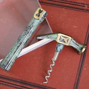 May include: Two antique bronze wine tools. One is a bottle stopper with a gold-coloured emblem. The other is a corkscrew with a silver worm and a gold-coloured emblem. Both tools have a green patina.