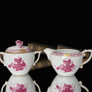 以下が含まれることがあります： ピンクの花柄と金色のアクセントが施された白い磁器製の砂糖入れとクリーマーのセット。セットは、ピンクとゴールドの繊細な花柄で飾られています。