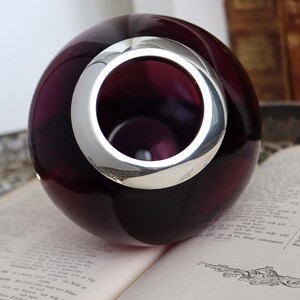 Peut inclure: Un vase sph&eacute;rique en verre violet fonc&eacute; avec un anneau argent&eacute; autour de l'ouverture. Le vase est pos&eacute; sur un livre ouvert. L'anneau argent&eacute; refl&egrave;te la lumi&egrave;re, cr&eacute;ant un contraste avec le verre sombre.