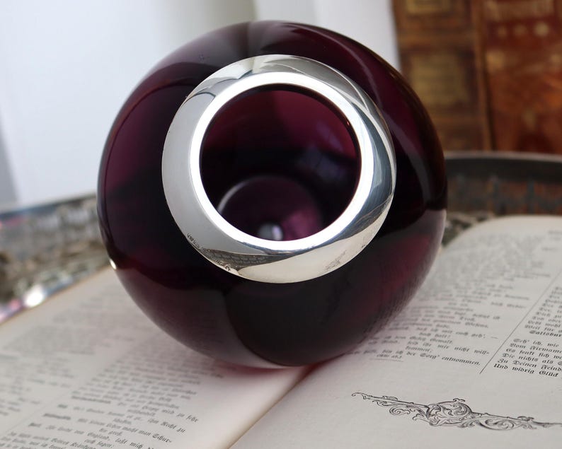 Peut inclure: Un vase sph&eacute;rique en verre violet fonc&eacute; avec un anneau argent&eacute; autour de l'ouverture. Le vase est pos&eacute; sur un livre ouvert. L'anneau argent&eacute; refl&egrave;te la lumi&egrave;re, cr&eacute;ant un contraste avec le verre sombre.
