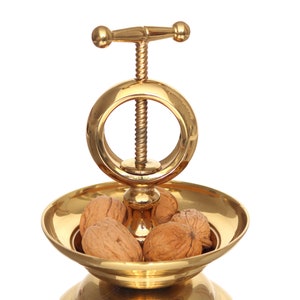 Puede incluir: Un cascanueces de metal dorado con una parte superior redonda abierta y un mecanismo de tornillo. El cascanueces tiene un pequeño plato debajo para contener nueces. Hay cuatro nueces en el plato.