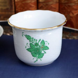 May include: White ceramic planter with a scalloped rim and gold trim. The planter features a green floral design on the front. The planter is a decorative item for displaying plants or flowers.