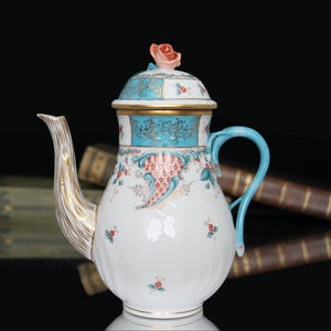 Op de afbeelding: Een witte porseleinen theepot met een goudomrande tuit, een lichtblauw handvat en een deksel bekroond met een gebeeldhouwde roze roos. Het lichaam is versierd met turquoise banden, goudfiligraan en kleine bloemmotieven in rood en groen.