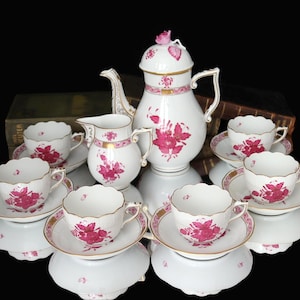 May include: A white porcelain tea set with a floral design in pink. The set includes a teapot, creamer, sugar bowl, and several teacups with saucers. Gold trim accents the edges of the cups and saucers.