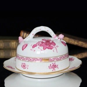 Puede incluir: Un mantequillero de porcelana blanca con un diseño floral rosa y un borde dorado. El mantequillero tiene una tapa abovedada con una manija y un plato a juego.