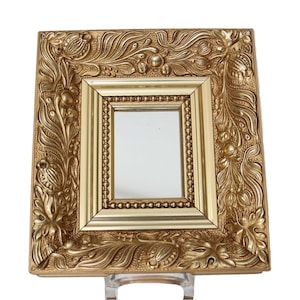 May include: A square, gold-colored picture frame with ornate, floral detailing. The frame has a smaller, rectangular opening in the center. The frame is supported by a clear stand, and the background is white.