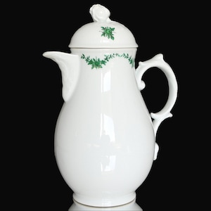 Könnte beinhalten: Weiße Porzellan-Kaffeekanne mit grünem Blumendesign und Deckel. Die Kanne hat einen geschwungenen Griff und einen runden Korpus.