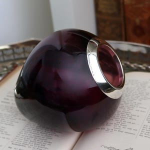 Peut inclure: Un objet en verre violet fonc&eacute; avec un bord argent&eacute;, pos&eacute; sur un livre ouvert. Le verre a une forme arrondie avec une surface lisse et r&eacute;fl&eacute;chissante. Le bord argent&eacute; ajoute un accent m&eacute;tallique contrastant.