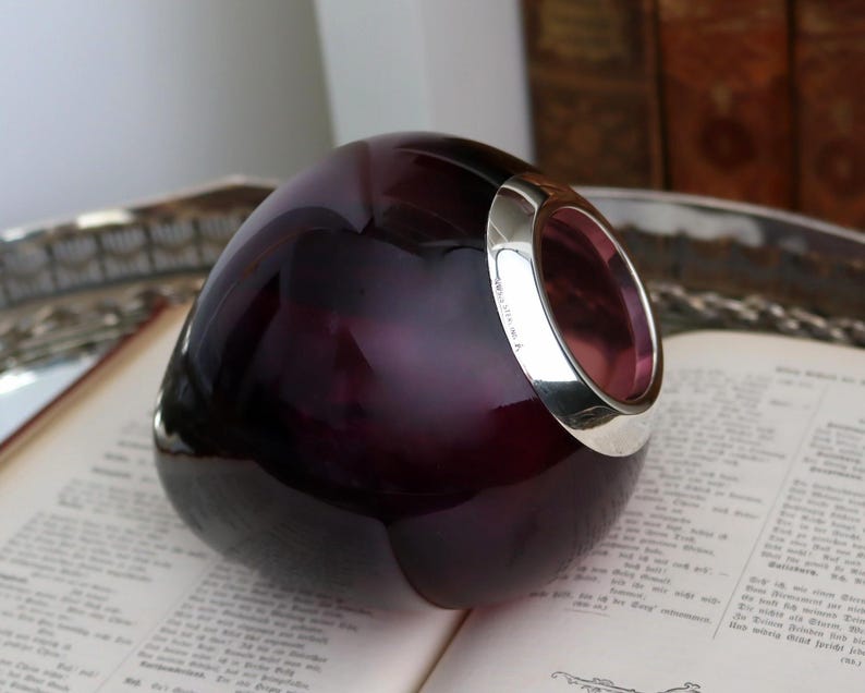 Peut inclure: Un objet en verre violet fonc&eacute; avec un bord argent&eacute;, pos&eacute; sur un livre ouvert. Le verre a une forme arrondie avec une surface lisse et r&eacute;fl&eacute;chissante. Le bord argent&eacute; ajoute un accent m&eacute;tallique contrastant.