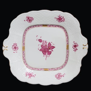 May include: A white porcelain serving plate with a square center and a raised, woven pattern around the edge. The plate has a pink floral design with gold accents.