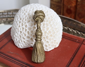 May include: A brass-colored tassel with a knotted top and braided details. The tassel is resting on a red book with black lines and a white, porous coral-like object in the background. Decorative home decor.