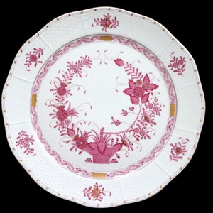 May include: A white porcelain dinner plate with a pink floral design and gold accents. The plate features a delicate floral pattern with a basket in the center.