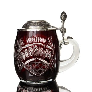 Puede incluir: Una jarra de cerveza de vidrio rojo rubí con intrincados diseños de vidrio tallado. La jarra tiene una tapa y un asa de metal plateado. El asa es de vidrio transparente. La tapa es abatible y tiene una parte superior decorativa.