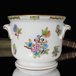 May include: White ceramic planter with colorful floral and butterfly designs. The planter has two handles and a gold rim. The floral design includes pink, blue, and orange flowers with green leaves. The butterflies are in various colors.