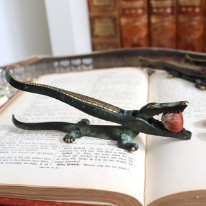 Pode incluir: Um quebra-nozes de bronze em forma de jacaré, com uma noz nas mandíbulas, repousando sobre um livro aberto. O quebra-nozes tem um corpo texturizado e um design detalhado. Várias nozes estão espalhadas por perto.