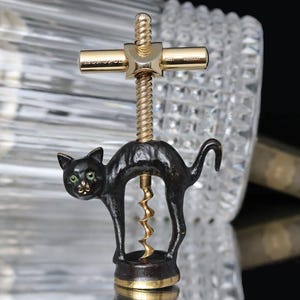 Puede incluir: Un sacacorchos con forma de gato negro con un tornillo y detalles dorados. El gato tiene ojos verdes y está sobre una base redonda. La parte superior del sacacorchos es dorada con el texto "WEST GERMANY".