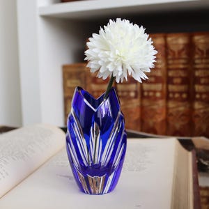 May include: A cobalt blue glass vase with a white flower. The vase has a unique, angular design with vertical gold accents. The flower is a large, white, spherical bloom. The vase is on an open book.