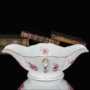 Op de afbeelding: Een witte porseleinen sauskom met een roze bloemenpatroon en gouden rand. De kom heeft een gegolfde rand en een handvat.