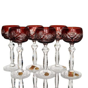 May include: Set of six ruby red crystal wine glasses. Each glass features a cut crystal design on the bowl and a clear, faceted stem. The glasses have a round base and are arranged against a white background.