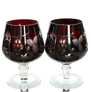 Puede incluir: Dos copas de brandy de cristal rojo rubí con diseños grabados de uvas y hojas. Las copas tienen un cuenco ancho, un tallo corto y una base circular. El cristal es transparente y los diseños grabados son visibles.