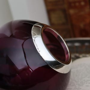 Peut inclure: Un vase en verre violet fonc&eacute; avec un bord argent&eacute;. Le vase a une forme arrondie avec une grande ouverture. Le bord argent&eacute; est marqu&eacute; "925 STERLING". L'arri&egrave;re-plan est flou.