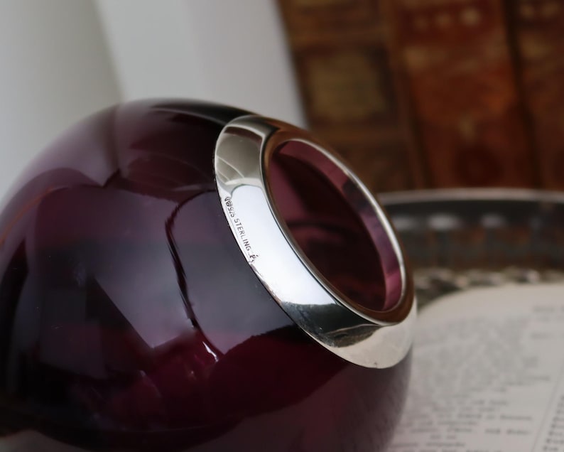 Peut inclure: Un vase en verre violet fonc&eacute; avec un bord argent&eacute;. Le vase a une forme arrondie avec une grande ouverture. Le bord argent&eacute; est marqu&eacute; "925 STERLING". L'arri&egrave;re-plan est flou.