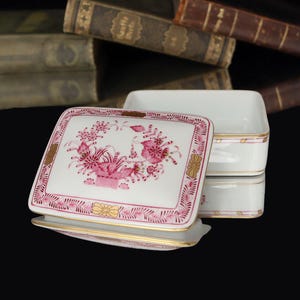 May include: A white porcelain trinket box with a rectangular shape and a matching lid. The lid features a floral design in shades of pink and gold accents. The box has a gold trim.