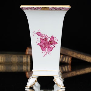 May include: A white porcelain vase with a pink floral design and gold trim. The vase has a square base with four feet and a rectangular body.