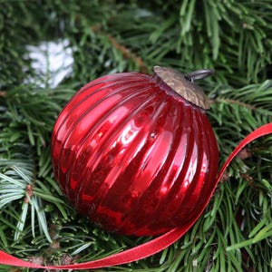 May include: A close-up of a red, ribbed glass Christmas bauble with a brass-coloured cap. The bauble rests on green pine needles, with a red ribbon weaving through the branches. Festive holiday decoration.