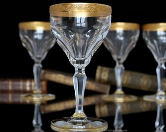 May include: Four clear glass stemmed drinking glasses with gold trim around the top rim. The glasses are arranged in a row on a black surface.