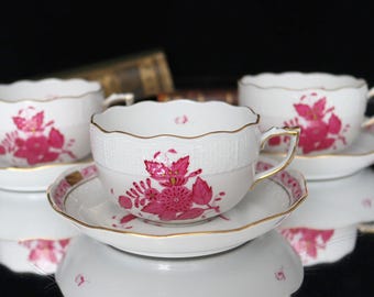May include: Three pink floral teacups and saucers with a white background and gold trim. The teacups have a scalloped edge and a delicate floral design.
