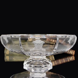 Puede incluir: Un cuenco de cristal transparente y facetado con base de pedestal. El cuenco presenta un emblema de cabeza de Medusa en el centro. El cuenco se utiliza probablemente con fines decorativos o de servicio.