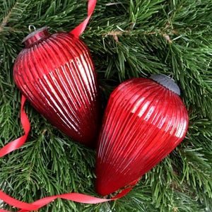 May include: Two red, ribbed glass Christmas ornaments with silver caps rest on a bed of green pine needles. A red ribbon weaves through the needles and around the ornaments, creating a festive holiday scene.